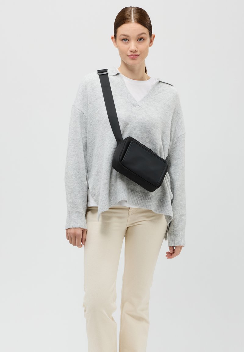 Schwarze Crossbody-Tasche aus glattem Material, rechteckige Form und Reißverschlussverschluss. Das Modell trägt einen hellgrauen Pullover und cremefarbene Hosen.
