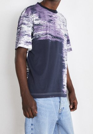 Mand iført en kortærmet t-shirt med lilla og hvidt abstrakt mønster på marineblå, kombineret med lyseblå denimjeans, mod en hvid baggrund.