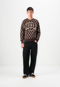 Young man standing wearing a brown patterned HUF sweater, loose black pants, and black sneakers with white laces against a white background.