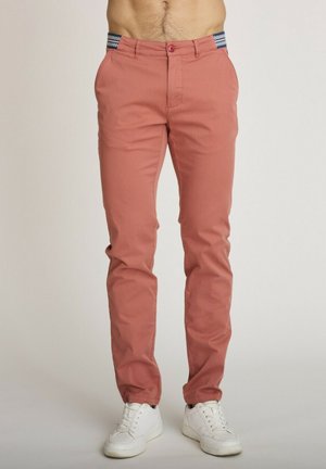 Homme portant un pantalon slim rose saumon avec poches latérales et des baskets blanches, visible de la taille aux pieds.