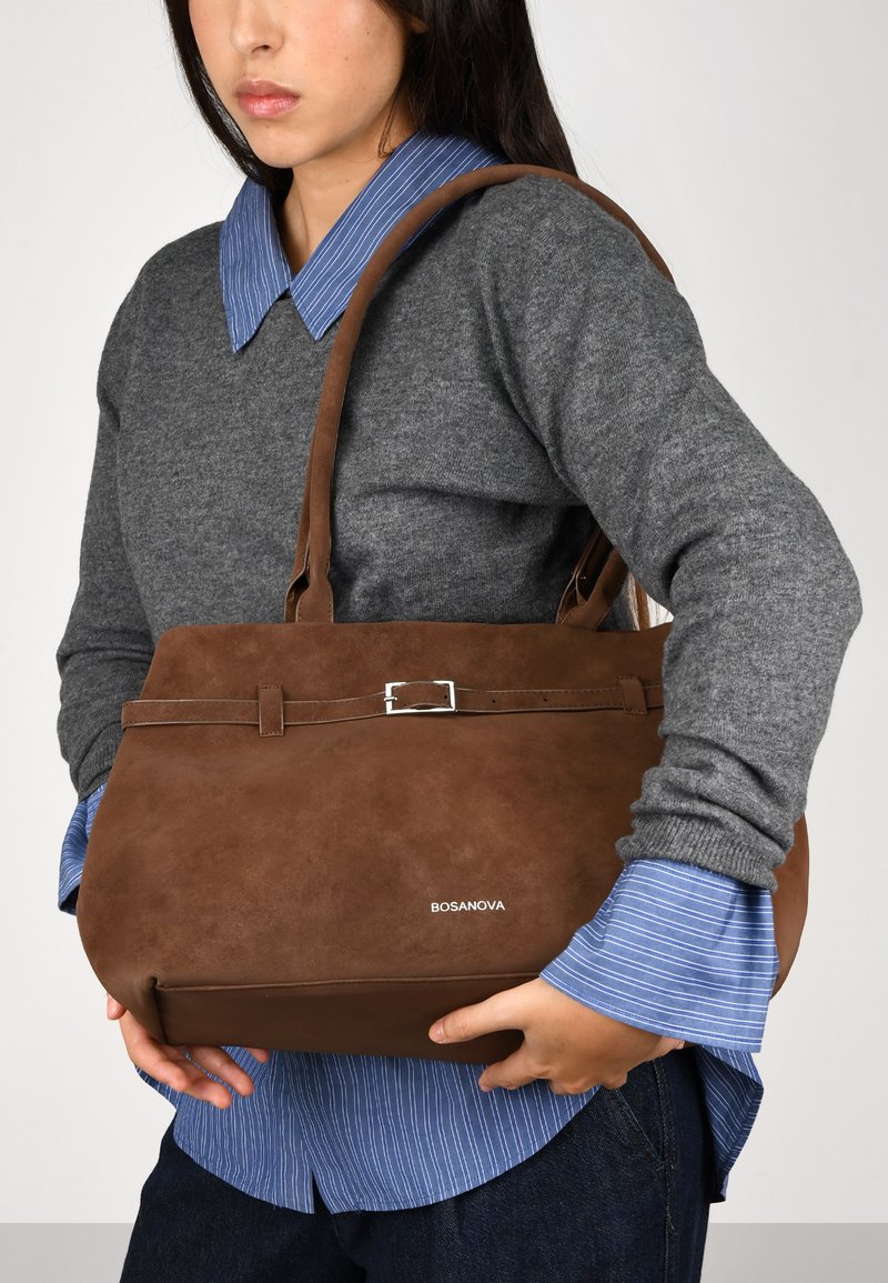 Bolso de mano de ante sintético marrón con forma plana, que presenta un detalle de hebilla plateada y dos asas. Logo "BOSANOVA" grabado en la parte frontal.