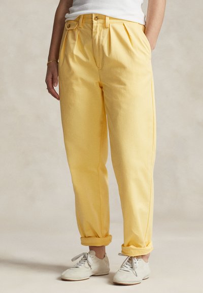 Gula bomullbyxor med veck, uppvikta muddar och knäppning med knapp. Bärs med vita sneakers mot en neutral bakgrund.