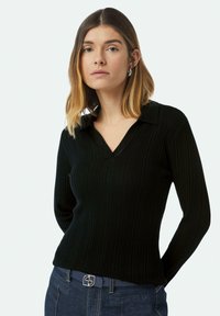 Schwarzes, geripptes Langarmoberteil mit V-Ausschnitt und flachem Kragen. Besitzt ein tailliertes Design und eine weiche Textur, kombiniert mit dunkelblauen Jeans.