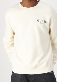 Felpa color crema realizzata in misto cotone, con scollatura a girocollo e un piccolo logo "GUESS JEANS" blu navy sul petto. Polsini a coste.