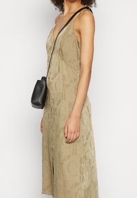 Beige bezrukávové šaty s texturovaným vzorem loga, tvar A-line a bočním rozparkem; doplněné malou černou crossbody taškou.