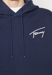 Marinblå bomullshoodie med dragsko på huvan, med en vit broderad logotyp på bröstet och ribbade muddar vid ärmarna.