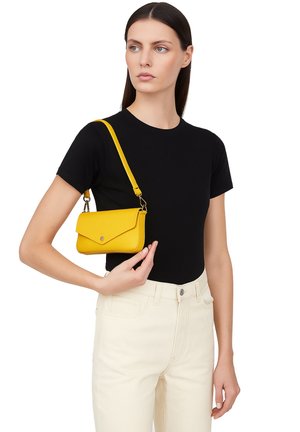 Sac à main en cuir texturé jaune avec un rabat, une bandoulière ajustable et un petit bouton métallique. Porté avec un t-shirt noir et un pantalon de couleur claire.