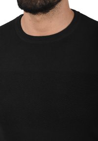Sort sweater med en glat tekstur og et kontrasterende strikkemønster over brystet, rund halsudskæring og ribbede ærmer.