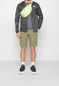 Lehká černá bunda, šedé tričko, olivově zelené kraťasy a černé tenisky s bílou podrážkou; viditelná jasně zelená ledvinka.