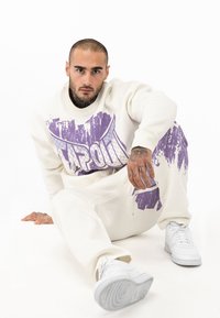 Hvid sweatshirt og sweatpants med lilla grafisk design, der har en struktureret print. Båret med hvide sneakers. Model sidder i positur.