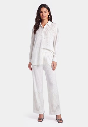 Donna con lunghi capelli scuri che indossa una camicia bianca con bottoni e pantaloni a gamba larga abbinati con piccoli motivi a puntini, in piedi con tacchi aperti.