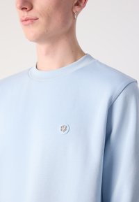 Svetlo modra kapuca s okroglo vratno odprtino, izdelana iz mehkega materiala. Opremljena z majhnim vezenim logotipom na levi strani prsnega koša.