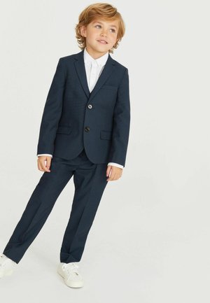 Abito blu navy con texture a motivo, giacca a un bottone e pantaloni dritti, abbinati a una camicia bianca e sneaker bianche.