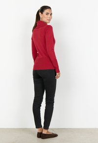 Kvinna med mörkt hår i en hästsvans, iklädd en röd polotröja, svarta slim-fit jeans och bruna ballerinas, stående mot en vit vägg.