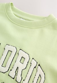 Sudadera verde claro con cuello redondo acanalado. Presenta un texto blanco audaz y peludo resaltado en negro. Textura suave en general.