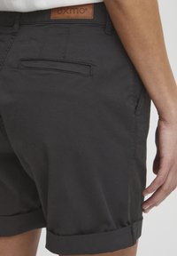Svarte shorts laget av lett materiale har rullet kant, sidelommer og en baklomme. En brun lærmerke viser merkenavnet.