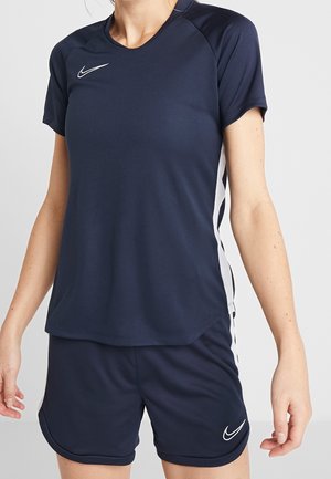 Kvinde iført navyblåt Nike sportssæt med hvidt logo, kortærmet top og matchende shorts med hvide sidelommer.