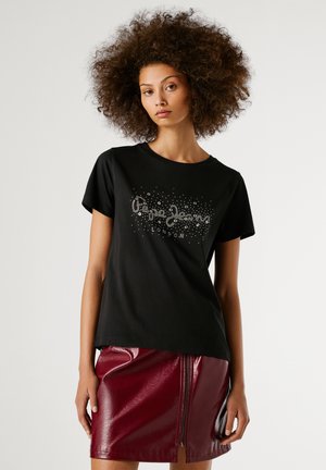 Jonge vrouw met volumineus krullend haar, gekleed in een zwart graphic t-shirt en een glanzende rode leren rok met een ritssluiting aan de voorkant, tegen een witte achtergrond.