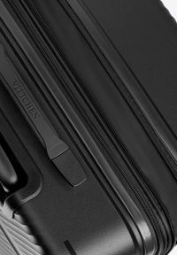 Černý pevný kufr s texturovaným povrchem, vybavený hladkým zipem a značkovou rukojetí. Design je elegantní a minimalistický.
