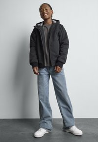 Μαύρο φερμουάρ μπουφάν με κουκούλα, γκρι μπλουζάκι, μπλε φαρδιά τζιν παντελόνια και λευκά αθλητικά παπούτσια. Λείο ύφασμα, άνετη εφαρμογή, καθημερινό στυλ.