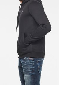 Svart bomullshuvtröja med kenguruficka, justerbar snörning i huvan, ribbade mudd och nederkant. I kombination med blå denimjeans.