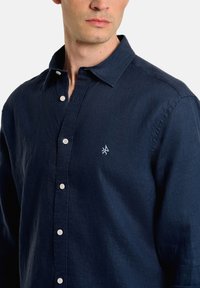 Camisa ajustada de color azul marino hecha de una tela texturizada, con cuello, botones blancos y un pequeño logo bordado en el pecho.