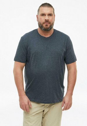 Man met baard en vastgebonden haar, met een donkergrijs T-shirt en beige broek, staande tegen een effen witte achtergrond.