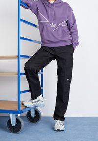 Lila hoodie med vit linje design, svarta byxor och ljusgrå skor med blå detaljer. Står bredvid en blå metallvagn på ett blått golv.