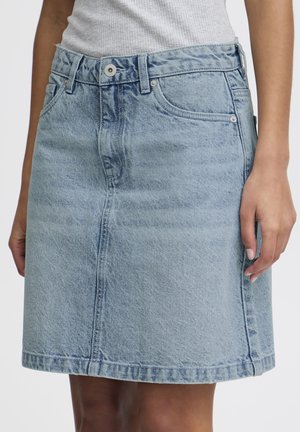 Jupe en denim bleu clair avec poches avant et fermeture par boutons, portée avec un débardeur blanc côtelé, dévoilant la taille jusqu'au milieu de la cuisse.