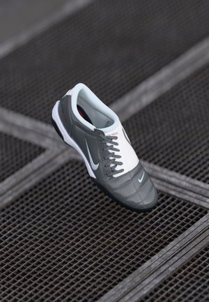 Grauer Sportschuh mit einer glatten Oberflächenstruktur, weißen Akzenten, auffälligen Schnürsenkeln und einer schwarzen Gummisohle, mit einem kleinen Logo an der Seite.