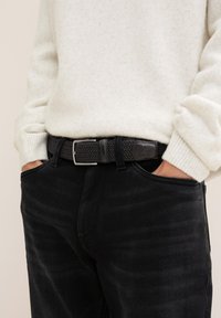Ceinture en cuir tissé noir avec une boucle rectangulaire en argent, portée sur un jean sombre et un pull texturé clair.