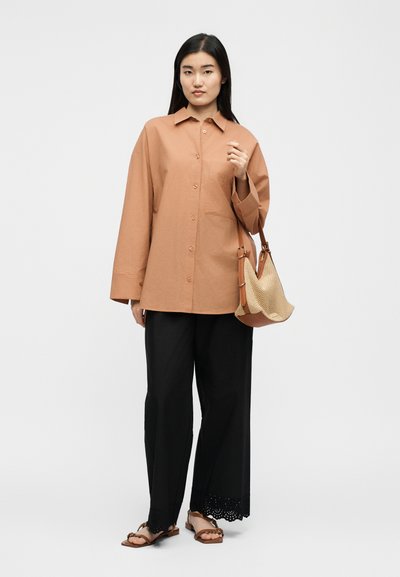 Γυναίκα που φοράει υπερμεγέθη μπεζ πουκάμισο με κουμπιά, μαύρο παντελόνι φαρδιάς γραμμής με δαντελένιο τελείωμα, καφέ σανδάλια, κρατώντας μπεζ πλεκτή τσάντα με δερμάτινα λουριά.