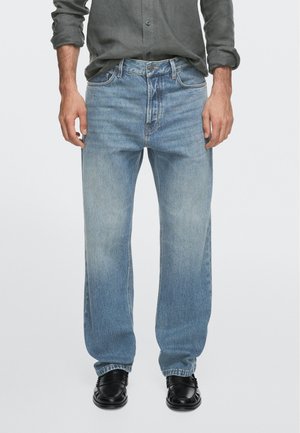 Uomo che indossa jeans azzurri sbiaditi a gamba dritta, scarpe nere e una camicia grigio scuro con bottoni e maniche arrotolate.