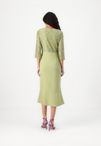 Robe verte avec des manches trois-quarts en dentelle, buste ajusté et ourlet évasé. Portée avec des talons strappy violets. Surfaces lisses et texturées.