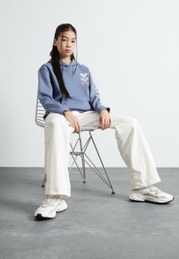 Felpa blu con dettagli grafiche sul petto e sulle maniche, abbinata a pantaloni cream a gamba larga e sneakers bianche. Il modello è seduto su una sedia di filo.