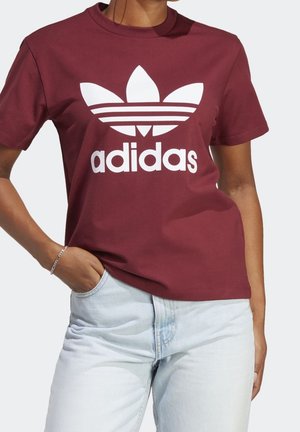 Personne portant un t-shirt Adidas bordeaux avec un logo blanc et un jean bleu clair, la main dans la poche, sur un fond clair uni.