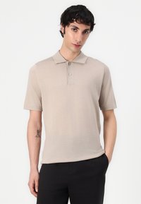 Polo beige avec une texture douce, à manches courtes et un col à trois boutons. Sans motifs, associé à un pantalon noir.