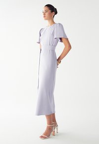 Svetlo-lavanda midi obleka s kratkimi napihnjenimi rokavi in pasom. Gladek material s tekočo silhueto, v kombinaciji z belimi sandali na peto.