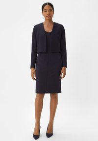 Marineblaues Kleid mit taillierter Silhouette und Rundhalsausschnitt, kombiniert mit einem kurz geschnittenen Blazer mit langen Ärmeln. Glatter Stoff und keine sichtbaren Muster.