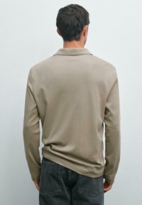 Man som står vänd bort med en beige långärmad skjorta och svarta jeans mot en enkel ljus väggbakgrund.