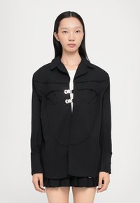 Camisa negra estructurada con bolsillos oversized, cierre de botones en el pecho y mangas largas con puños abotonados. Textura de tela suave.