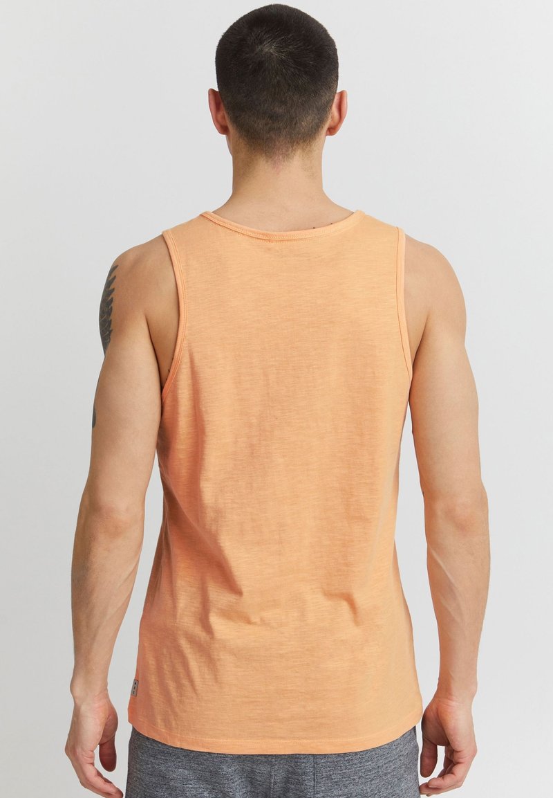 Homme aux cheveux courts et foncés portant un t-shirt orange sans manches et un short gris, vu de dos sur un fond uni.