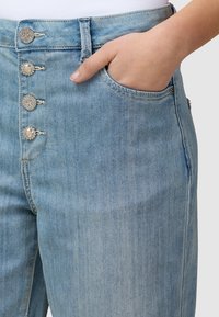 Ruka spočívá v kapse světle modrých džín s dekorativními perlami a kovovými knoflíky na pasku.