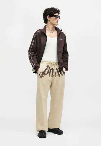 Άνδρας που φοράει μαύρα γυαλιά ηλίου, καφέ μπουφάν με φερμουάρ, λευκό φανελάκι, μπεζ παντελόνι με σκούρα γραφικά κείμενα και μαύρα αθλητικά παπούτσια, με τα χέρια στις τσέπες, κοιτάζοντας δεξιά.