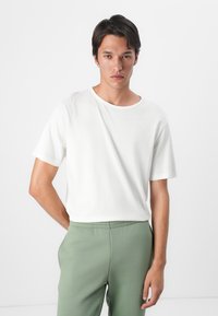Hvid bomulds T-shirt med korte ærmer, rund halsudskæring, båret med grønne sweatpants. Glat tekstur, afslappet pasform, ensartet design.
