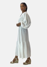 Robe longue blanche avec des manches bouffantes et un ourlet à volants. Portée avec des sandales à talons compensés dorées. Tissu lisse et coupe décontractée.