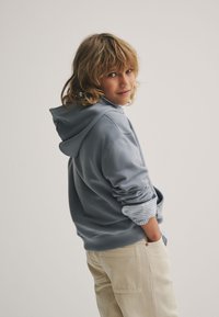 Heleroheline kapuutsiga dressipluus, mille lõige on lõdva istuvusega ning mille varrukatest paistavad triibulised mansetid. Kombineeritud kreemika värvi pükstega.