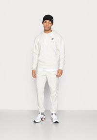 Un hoodie gris clair et un pantalon de survêtement en tissu doux, avec un petit logo noir, associés à des baskets blanches et un béret noir.