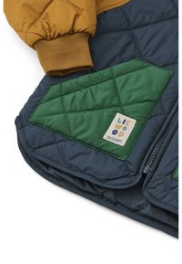 Quilted jacket featuring a mustard yellow upper, navy blue body, and green diamond-patterned pockets with a brand label. Soft texture, zipper closure.