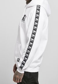 Uomo che indossa un hoodie bianco di Starter con strisce nere sul collo e sulla tasca frontale, in piedi di lato contro uno sfondo uniforme.